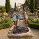 Large Traditional Dancing Girls Bronze Sculpture by Giant Sculptures, three barefoot girls circling a flower stem, displayed on a stone plinth in a formal garden.