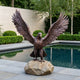 Large Traditional Eagle Bronze Sculpture by Giant Sculptures, wings fully spread, perched on a stone boulder beside a reflecting pool with hedges behind.