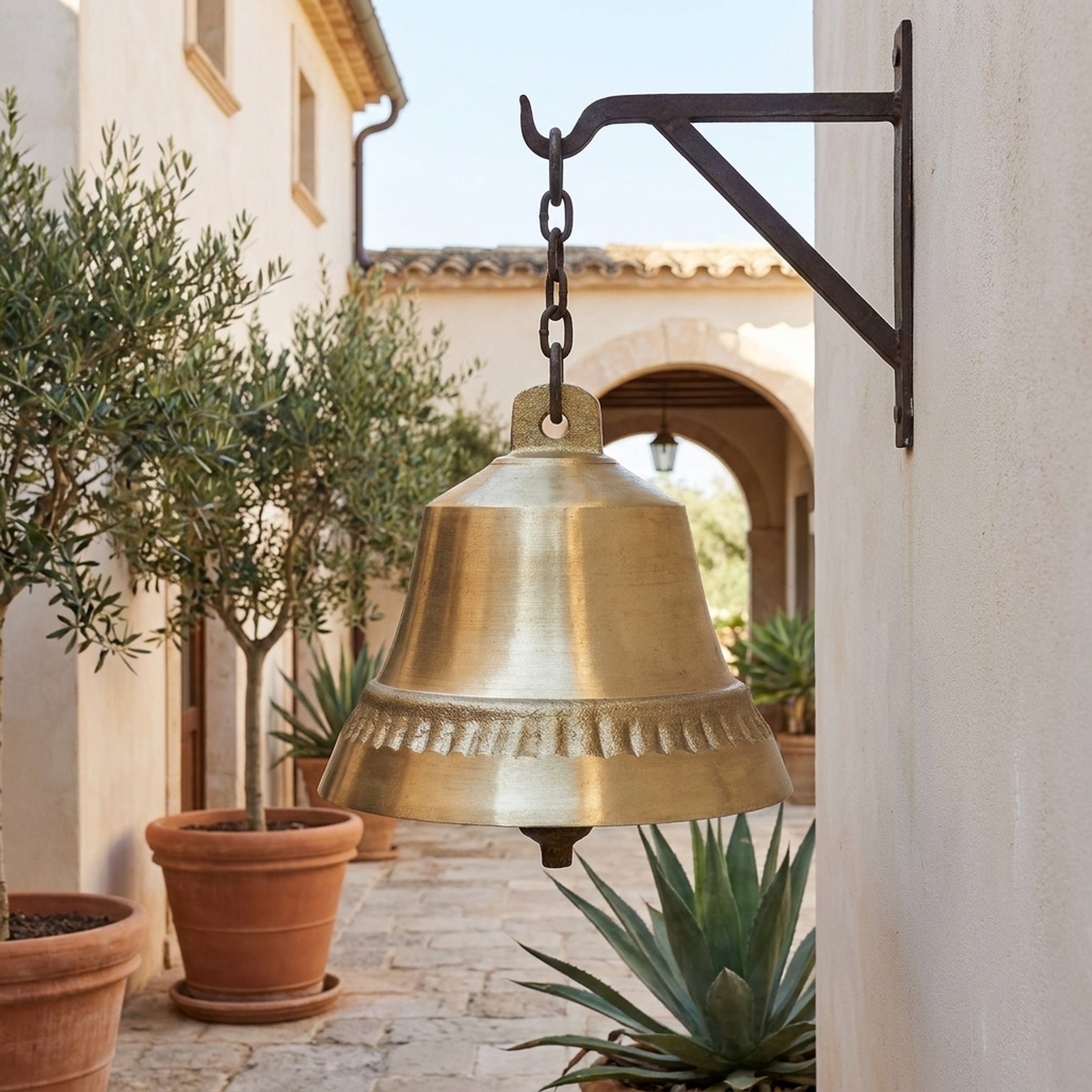 Large Traditional Hanging Bell Bronze Sculpture by Giant Sculptures suspended from an iron wall bracket in a Mediterranean courtyard with olive trees and terracotta pots.