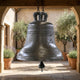 Large Traditional Inscribed Church Bell Bronze Sculpture by Giant Sculptures, 120cm, suspended by chains in an archway courtyard with olive trees and lavender.