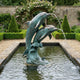 Large Traditional Leaping Dolphins Bronze Fountain by Giant Sculptures, two verdigris dolphins arcing from a sculpted wave, spouting water into a walled garden pool.
