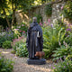 Large Traditional Robed Saint Bronze Statue 210cm by Giant Sculptures, holding a cross-staff and book with a reddish-brown pig at his feet, in a flowering garden.
