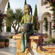 Large Traditional Seated Girl Bronze Sculpture by Giant Sculptures with green-brown verdigris patina, seated on a stone wall holding a ball in a Mediterranean courtyard.