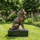 Large Traditional Seated Husky Dog Bronze Sculpture by Giant Sculptures, side profile on a dark granite plinth in a garden with flowerbeds and trees.