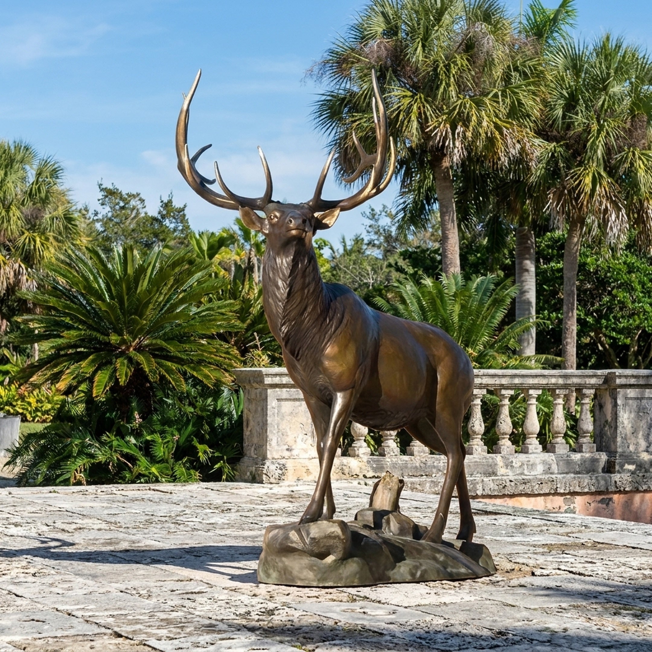 Life-Size Alert Bull Elk Bronze Sculpture by Giant Sculptures shown on a stone terrace beside palms and a balustrade, head raised with wide antlers.