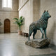 Life-Size Alert Husky Dog Bronze Sculpture by Giant Sculptures with verdigris patina on a boulder base, displayed in a stone-walled hallway near a potted palm.