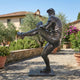 Life-Size Baseball Pitcher Bronze Statue by Giant Sculptures with dark patina, shown mid wind-up on a stone patio beside a Tuscan villa with olive trees.