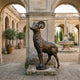 Life-Size Bighorn Ram Bronze Sculpture by Giant Sculptures in warm brown patina, displayed in an arched stone courtyard near a fountain and potted olive trees.