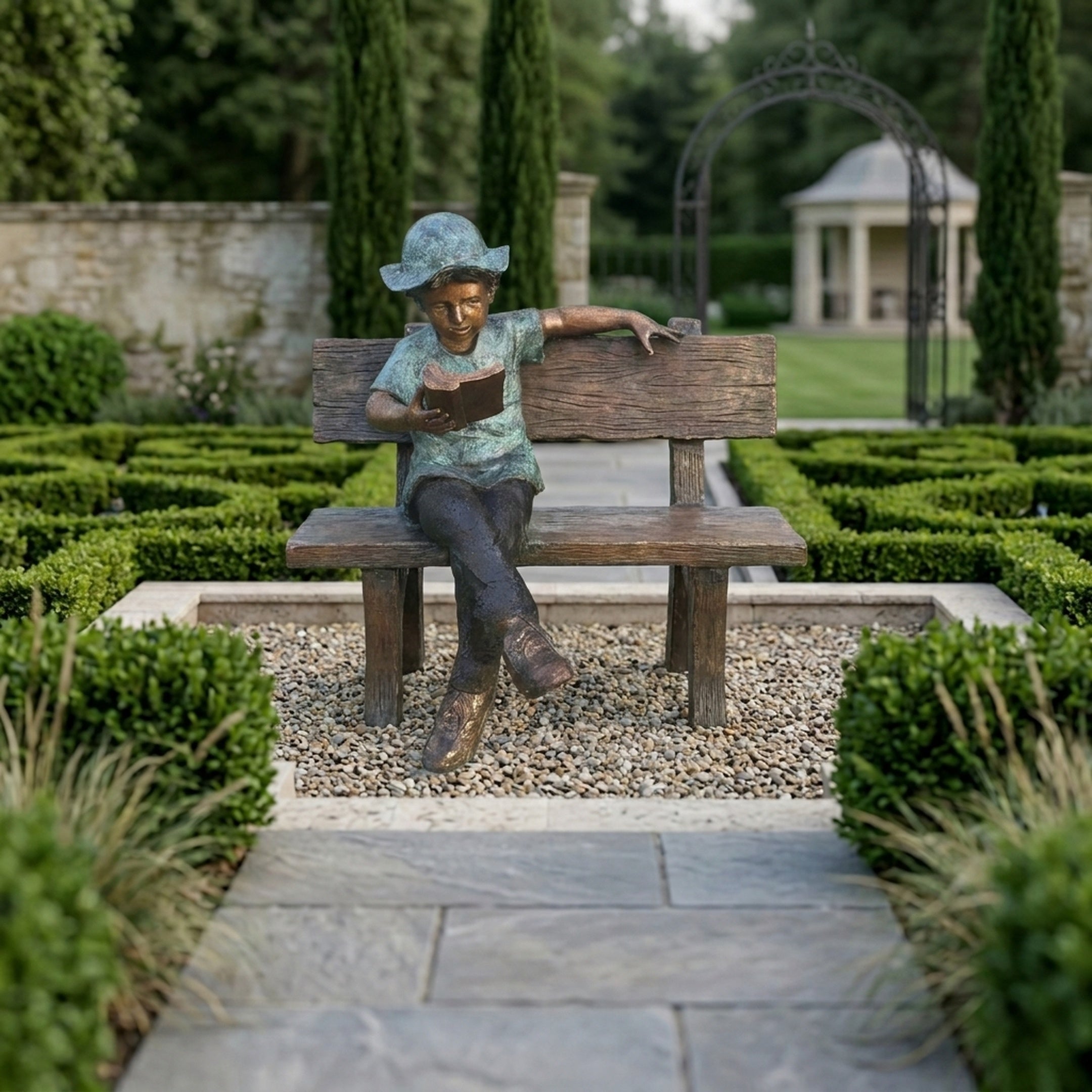 Giant Sculptures Life-Size Boy Reading on Bench Bronze Sculpture 150cm with green verdigris and brown patina, displayed on gravel in a formal hedged garden.