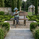Giant Sculptures Life-Size Boy Reading on Bench Bronze Sculpture 150cm with green verdigris and brown patina, displayed on gravel in a formal hedged garden.