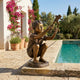 Life-Size Boy with Jumping Frog Bronze Sculpture by Giant Sculptures, crouching with frog held aloft, displayed on a sunlit stone poolside terrace.