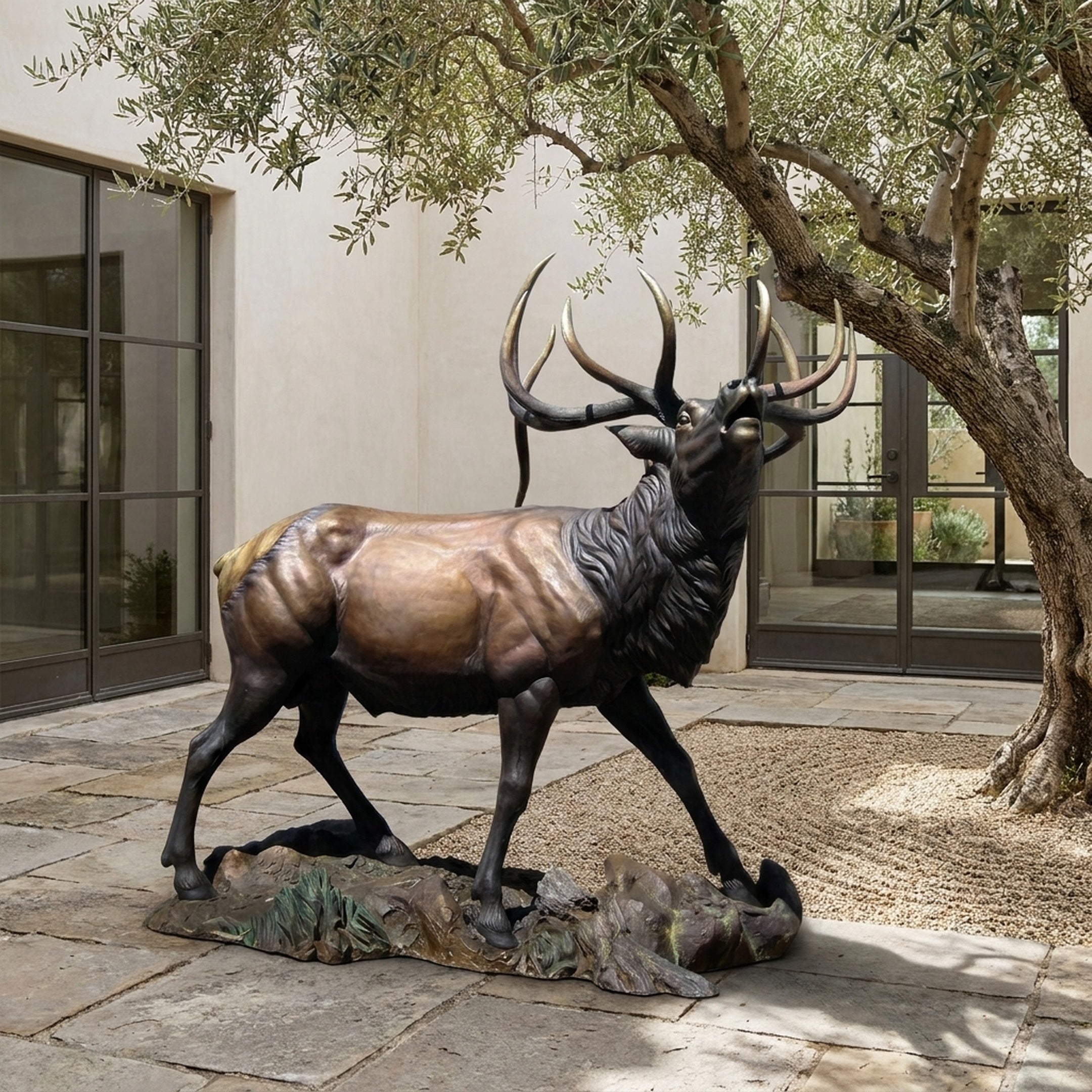 Life-Size Bugling Bull Elk Bronze Sculpture by Giant Sculptures shown mid-stride with head raised and antlers swept back, displayed in a stone courtyard beside an olive tree.