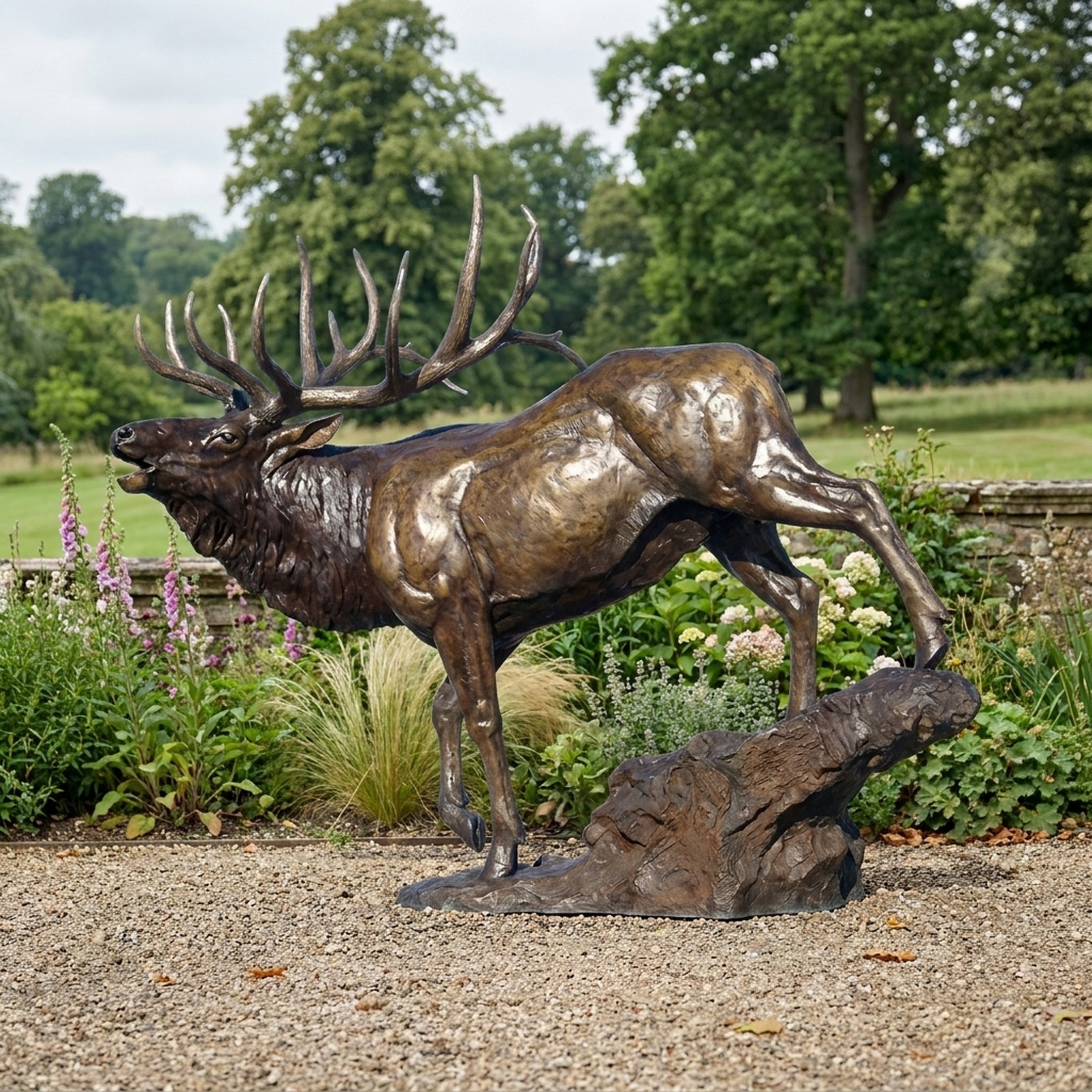 Life-Size Bull Elk Striding Bronze Sculpture by Giant Sculptures shown in side profile with head raised mid-bugle, displayed on a gravel garden path beside flowering borders.
