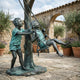 Life-Size Children on Swing Bronze Sculpture by Giant Sculptures, verdigris-patinated boy pushing a girl on a rope swing tied to a tree, in a stone courtyard.