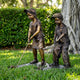 Life-Size Children Playing Golf Bronze Sculpture by Giant Sculptures, showing a girl and boy in caps gripping clubs over balls, set on grass beside a hedge.
