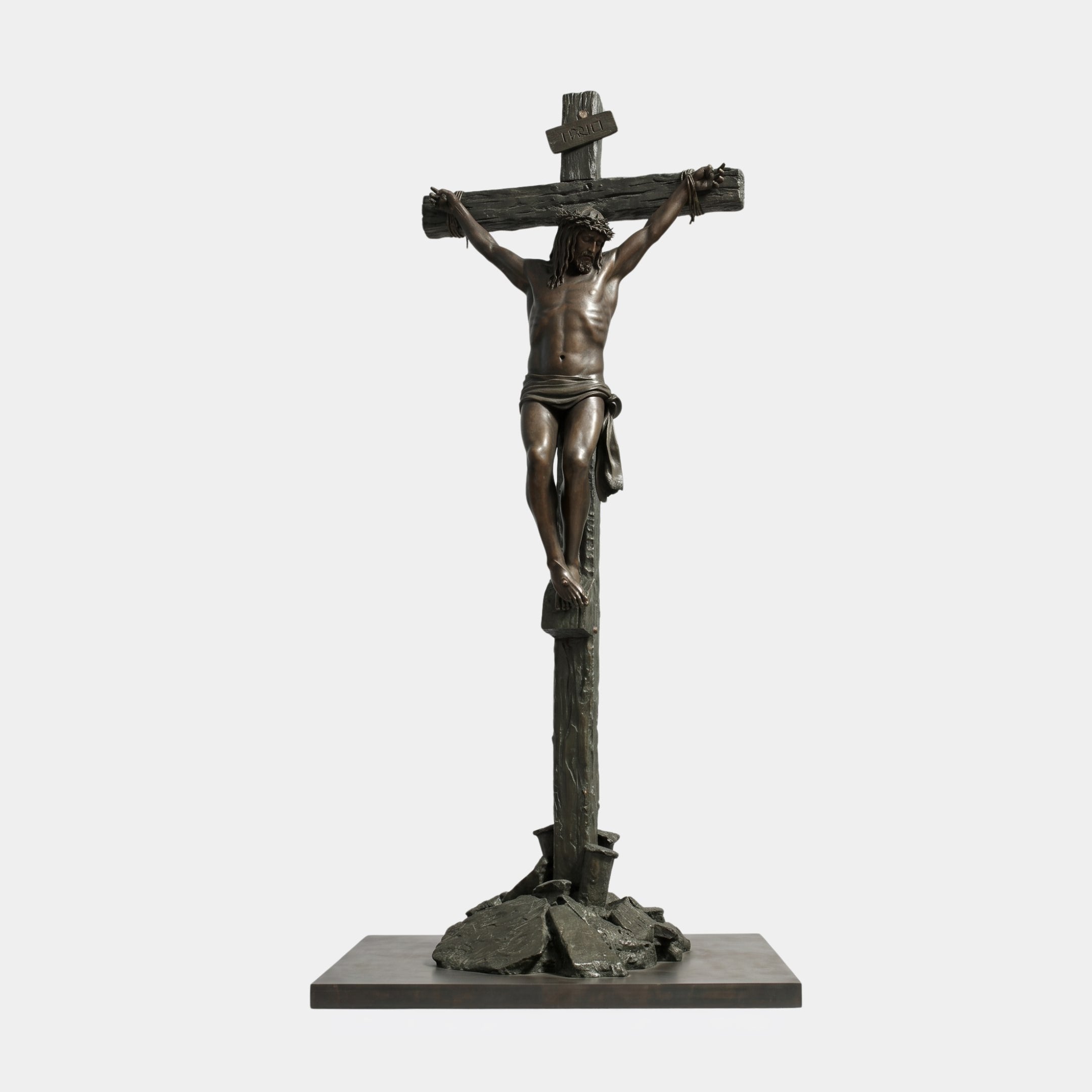 Life-Size Classical Crucifixion of Christ Bronze Sculpture by Giant Sculptures, dark patinated figure on a rugged cross with rocky base, plain white background.