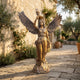 Life-Size Classical Female Angel Bronze Sculpture by Giant Sculptures with raised torch and spread wings, displayed on a stone courtyard beside olive trees.