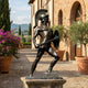 Life-Size Classical Greek Warrior Bronze Statue by Giant Sculptures in dark patina, sword and shield raised in combat stance, displayed on a Tuscan terrace.