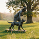 Life-Size Classical Hunter and Wolf Bronze Sculpture by Giant Sculptures, dark patina figure striding beside a wolf, displayed on grass beneath a large tree.