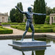 Life-Size Classical Nude Male Deity Bronze Sculpture by Giant Sculptures in throwing pose on a granite plinth in a formal garden pond with topiary.