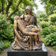 Life-Size Classical Pietà Bronze Sculpture by Giant Sculptures, 180cm, displayed on a dark granite plinth in a lush garden with ferns and flowering shrubs.