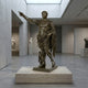 Life-Size Classical Roman Commander Bronze Statue by Giant Sculptures, 150cm dark-brown figure in cuirass with arm raised, on a travertine plinth in a gallery.