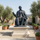 Giant Sculptures Life-Size Classical Seated Figure Bronze Statue on a stone plinth in a Mediterranean courtyard with olive trees, lavender, and potted shrubs.