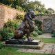 Life-Size Classical Seated Lion Bronze Sculpture by Giant Sculptures, warm brown patina, on a low plinth in a walled garden with a timber gate.