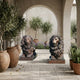 Life-Size Classical Seated Lions Bronze Sculpture by Giant Sculptures displayed as a mirrored pair on a stone terrace with olive trees, terracotta pots, and arched stucco walls.