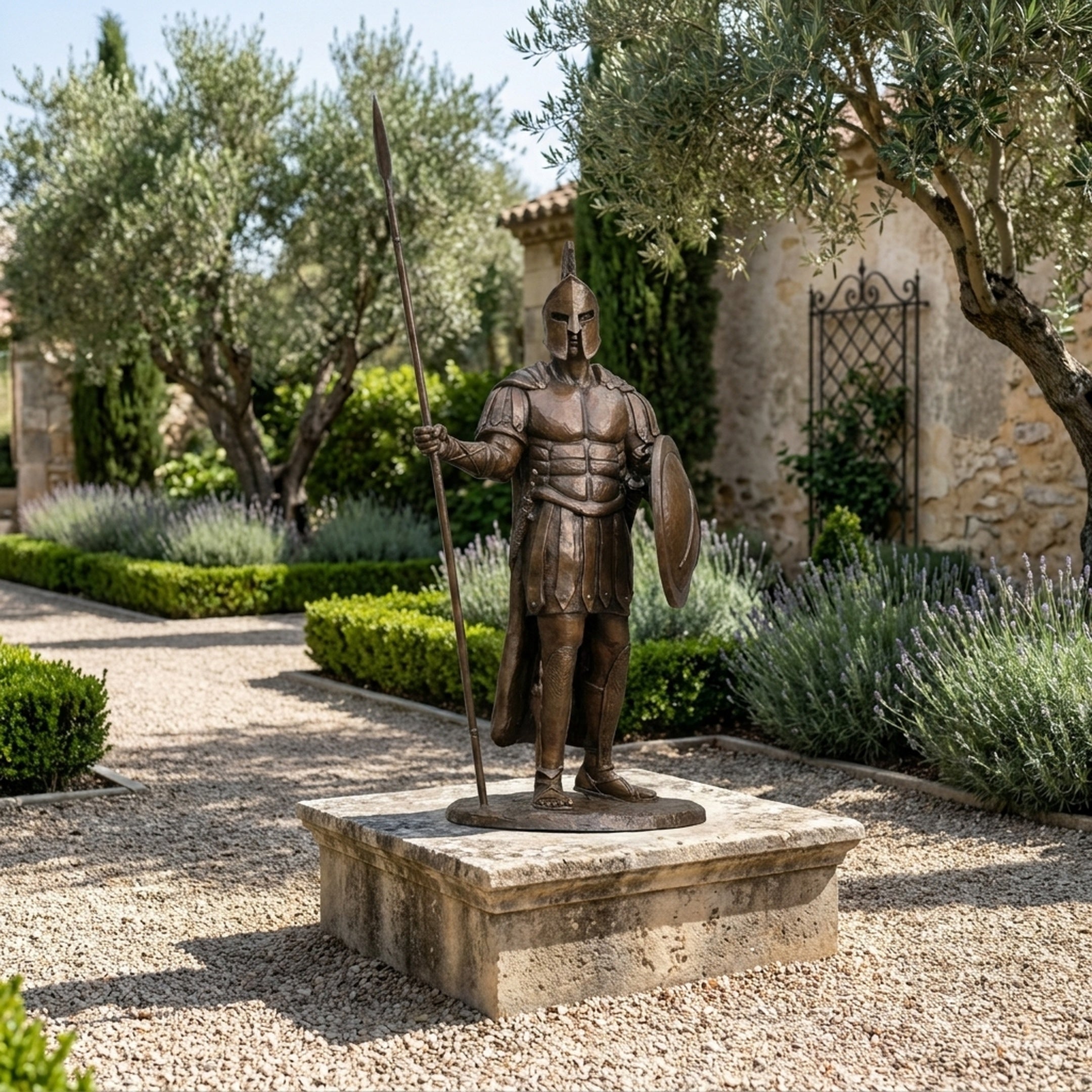 Life-Size Classical Spartan Warrior Bronze Statue by Giant Sculptures on a stone plinth in a Mediterranean garden with lavender, olive trees, and stone buildings.