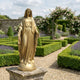 Life-Size Classical Virgin Mary Bronze Statue in gold finish by Giant Sculptures, displayed on a stone plinth in a formal lavender and rose garden.