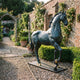 Life-Size Classical Walking Horse Bronze Sculpture by Giant Sculptures with dark patina and gold hooves, displayed on gravel in a walled rose garden.