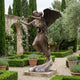 Life-Size Classical Winged Angel Bronze Sculpture by Giant Sculptures, shown holding a child aloft in a formal garden with stone arch and lavender borders.