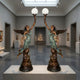 Life-Size Classical Winged Angel Pair Floor Lamp Bronze Sculpture by Giant Sculptures, mirrored figures with verdigris drapery raising frosted globe lamps in a gallery.