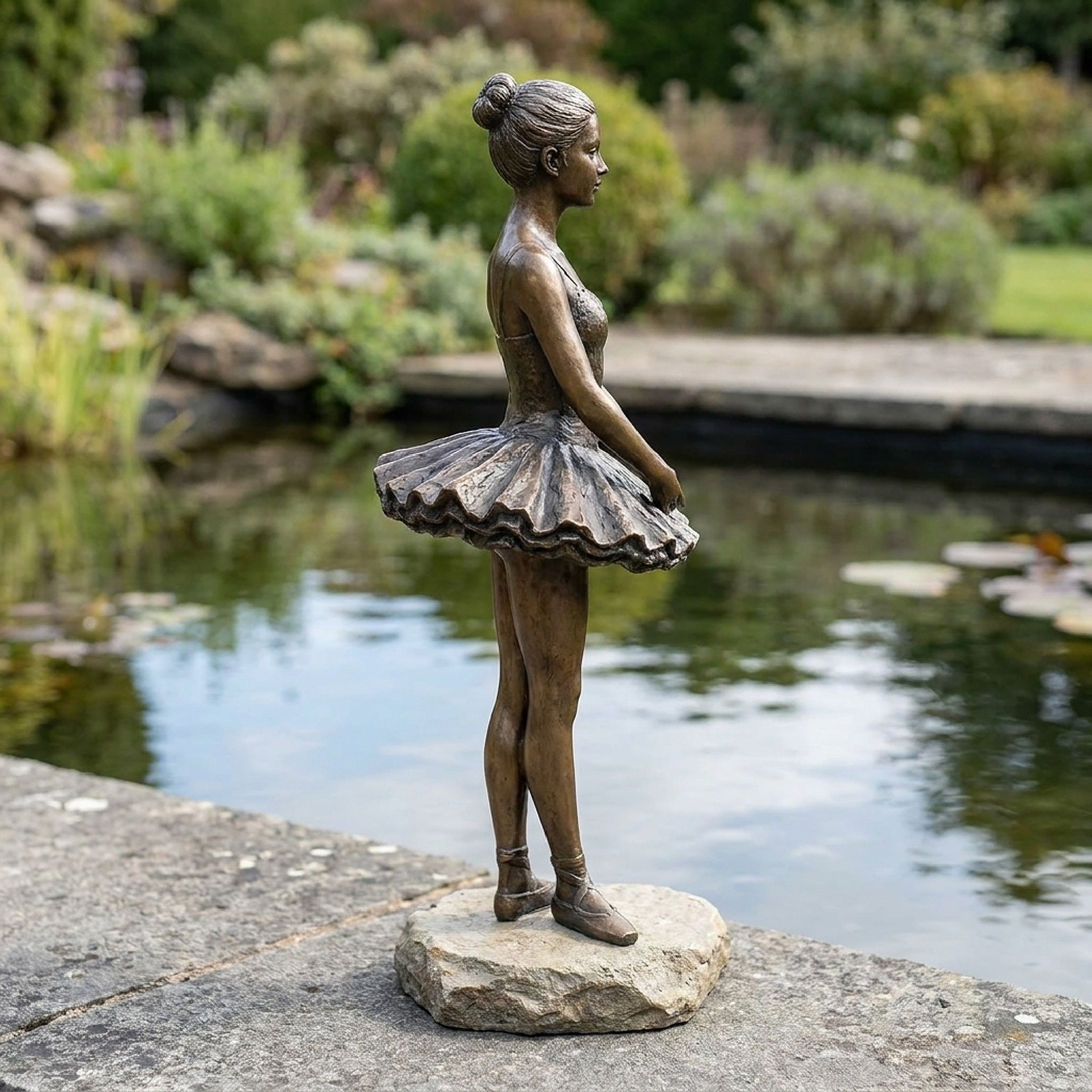 Life-Size Contemporary Ballerina Bronze Sculpture by Giant Sculptures, side profile on a stone base beside a garden pond with greenery behind.