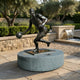 Life-Size Contemporary Basketball Player Bronze Sculpture by Giant Sculptures, mid-dribble pose on a granite plinth in a sunlit garden beside a water feature.