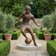 Life-Size Contemporary Footballer Dribbling Bronze Sculpture by Giant Sculptures shown mid-stride with the ball on a round stone base in a hedged garden.