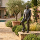 Life-Size Contemporary Male Figure Bronze Sculpture by Giant Sculptures, suited man with foot on a football, displayed on a stone plinth in a Mediterranean garden.