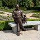 Life-Size Contemporary Seated Man and Cat Bronze Sculpture by Giant Sculptures, shown on a granite bench beside manicured garden hedges and gravel paths.