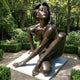 Life-Size Contemporary Seated Nude Female Bronze Sculpture by Giant Sculptures, warm brown patina figure seated on a stone plinth in a leafy garden setting.