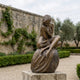 Life-Size Contemporary Seated Nude Female Figure Bronze Sculpture by Giant Sculptures, shown on a stone plinth in a walled garden with hedging and olive trees.
