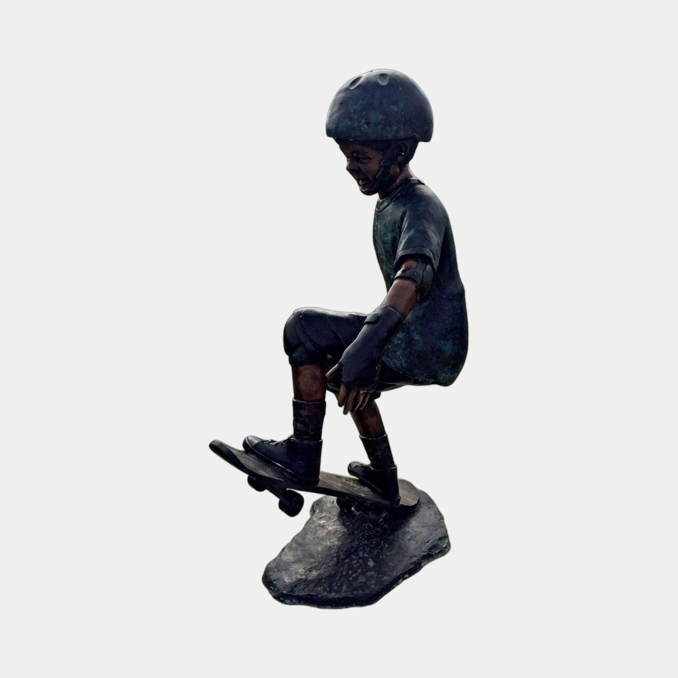 Life-Size Contemporary Skateboarding Boy Bronze Sculpture by Giant Sculptures, side view of a helmeted boy crouched mid-trick on a skateboard atop a rocky base.