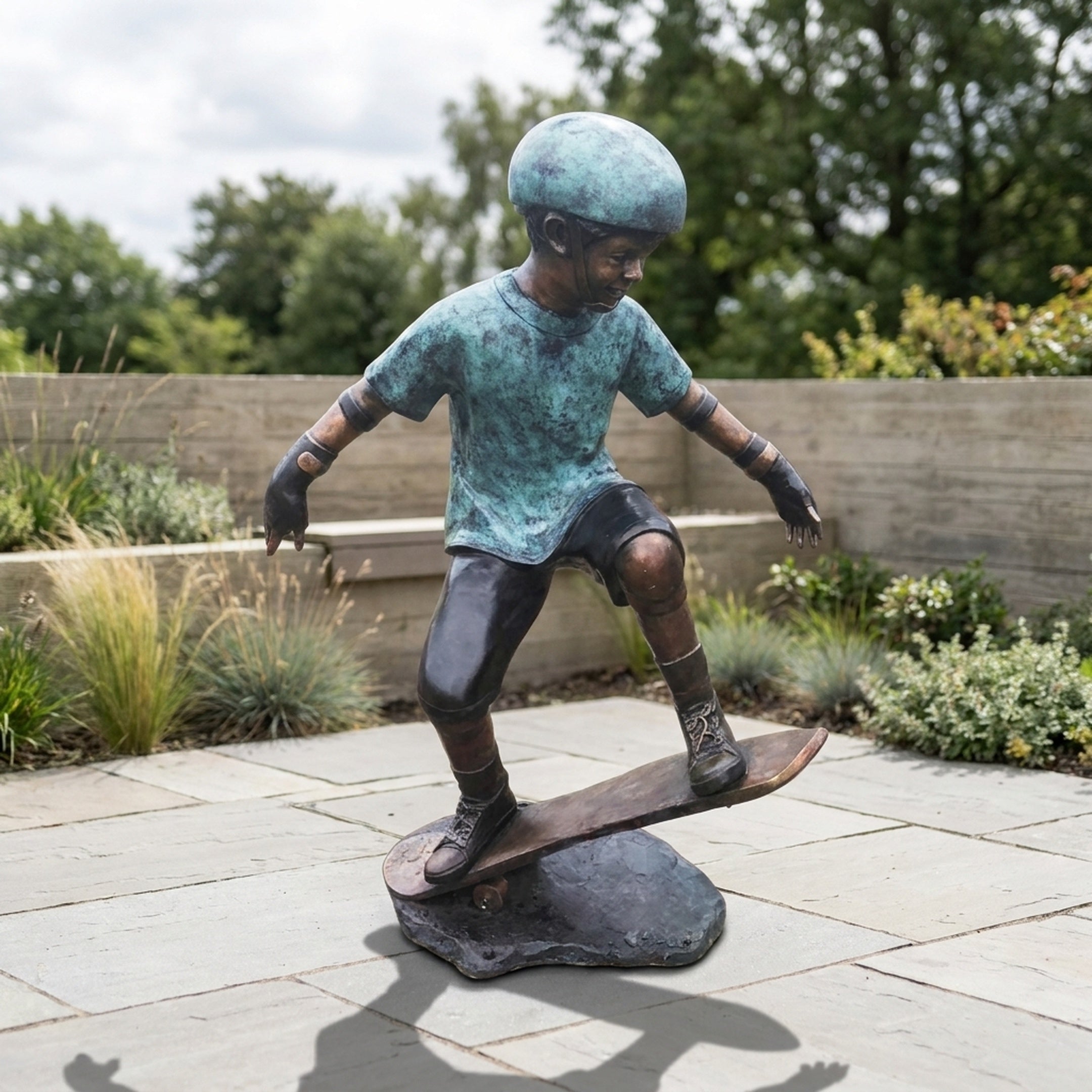 Life-Size Contemporary Skateboarding Boy Bronze Sculpture by Giant Sculptures, arms spread riding the board with teal verdigris top and dark legs, on a garden patio.