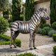 Life-Size Contemporary Standing Zebra Bronze Sculpture by Giant Sculptures shown side-on with black and golden-tan stripes, displayed on gravel in a Mediterranean garden.
