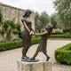 Life-Size Contemporary Two Girls Bronze Sculpture by Giant Sculptures on a stone plinth in a hedged garden, depicting a taller girl and child holding hands mid-play.