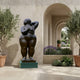 Life-Size Contemporary Voluptuous Female Figure Bronze Sculpture by Giant Sculptures on a green plinth in a stone courtyard with olive trees and arched passage.