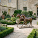 Life-Size Galloping Horse Pair Bronze Sculpture by Giant Sculptures displayed on a stone courtyard path beside boxwood hedges and a weathered stone manor.