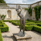 Life-Size Golfer Mid-Swing Bronze Sculpture by Giant Sculptures shown in follow-through pose on a stone plinth in a formal hedged garden with balustrade.
