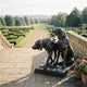 Life-Size Hunting Dogs Bronze Sculpture by Giant Sculptures on a stone terrace overlooking a formal hedge garden, with one hound standing and one seated.