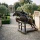Life-Size Jockey and Horse Clearing a Jump Bronze Sculpture by Giant Sculptures on castors in a formal garden with olive trees and a stone pavilion.
